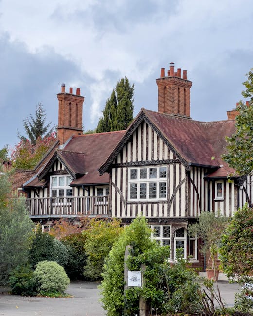 A traditional Tudor-style house featuring black timber framing and white infill panels, set amidst a lush garden with a variety of plants and trees. The garden includes trimmed bushes, flowering shrub