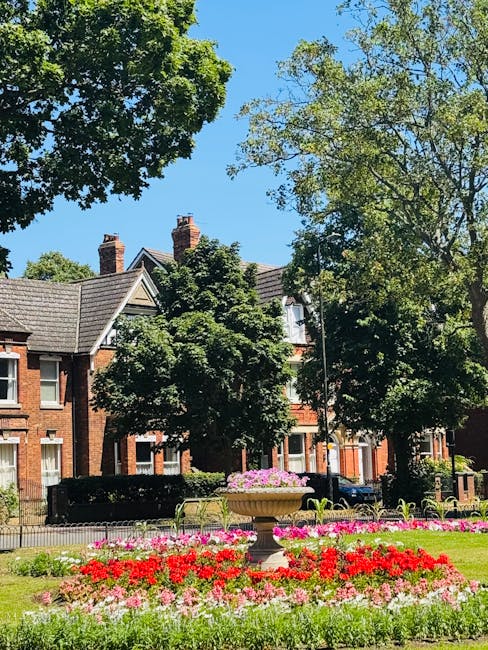A well-maintained urban garden featuring vibrant flower beds filled with red, pink, and white blooms arranged in neat, curved borders around a central stone planter filled with pink flowering plants. 
